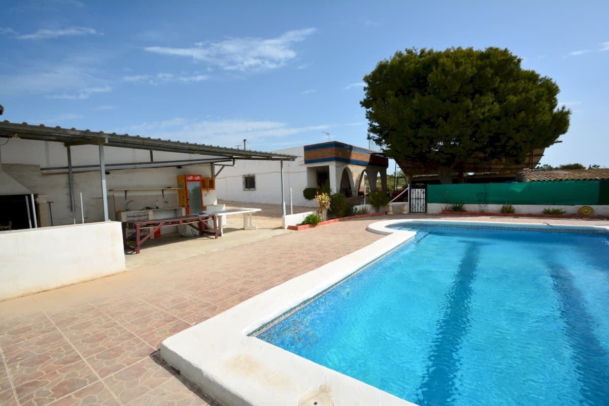Nestled in the emerging residential sector of El Mirador, Guardamar del Segura, this property offers a unique opportunity to own a substantial private estate within a rapidly developing area. The expansive 2,930 m² plot hosts a single-level home with a generous 311 m² of constructed area, classified as urban property and fully serviced with water and electricity, soon to be connected to a central sewage system.

The main house features a practical layout with four bedrooms, two bathrooms, a separate kitchen, and a cozy living room complete with a fireplace. A rooftop terrace enhances the space, providing views of the surrounding area. Adjacent to the main home, a guest house offers two additional bedrooms, a kitchen, and a full bathroom, ensuring privacy with its own entrance. The property also includes a closed garage with ample storage, multiple sheds, covered parking, and a summer BBQ area, making it ideal for both residential and leisure purposes. A large swimming pool is a centerpiece of the garden, promising endless relaxation.

While the property requires renovation, it presents a fully legal investment opportunity, with the possibility to rebuild entirely within the current footprint of 311 m². Conveniently located, it is a short drive from Ciudad Quesada and Guardamar del Segura, with amenities such as a supermarket just five minutes away. Alicante Airport is easily accessible within 40 minutes, ensuring excellent connectivity.

This property is an exceptional prospect for those seeking a spacious home or investment in a tranquil yet accessible location. Visits are available upon request. Taxes and associated purchase expenses are not included in the price.