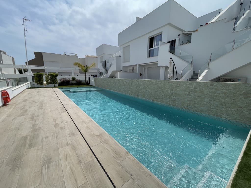 Stylish Ground-Floor Bungalow with Private Terrace and Communal Pool AccessThis beautifully presented ground-floor bungalow offers an inviting blend of contemporary design, comfort, and outdoor living — ideal as a permanent residence, holiday retreat, or rental investment.The home opens into a bright and airy open-concept living space, where modern aesthetics meet practicality. The fully fitted kitchen features clean white lines, integrated appliances, and a functional island with breakfast seating — perfect for cooking and entertaining alike. The adjacent living area is tastefully furnished and flows seamlessly out to the private terrace, enhancing the indoor-outdoor lifestyle.Step outside to a spacious, sunny terrace, where there’s ample room for dining, lounging, and enjoying the peaceful surroundings. Thoughtful touches like decorative wall art, potted greenery, and shaded seating areas create a relaxed Mediterranean atmosphere.The property includes two generous bedrooms with built-in wardrobes. The master bedroom features a warm, welcoming design and benefits from a private en suite bathroom, adding an extra layer of comfort and privacy. The second bedroom — currently arranged with twin beds — is perfect for guests or family.There are two modern bathrooms in total, both designed with high-quality finishes, spacious walk-in showers, and stylish vanities — with the en suite offering additional convenience for the main bedroom.A discreet utility closet houses the washing machine, water heater, and cleaning supplies, keeping the outdoor space tidy and functional.This property is part of a secure, well-maintained residential community with access to a sleek communal pool surrounded by lush greenery — an ideal setting for relaxing and cooling off.This is a turn-key home that perfectly balances convenience, style, and comfort — just bring your suitcase.
