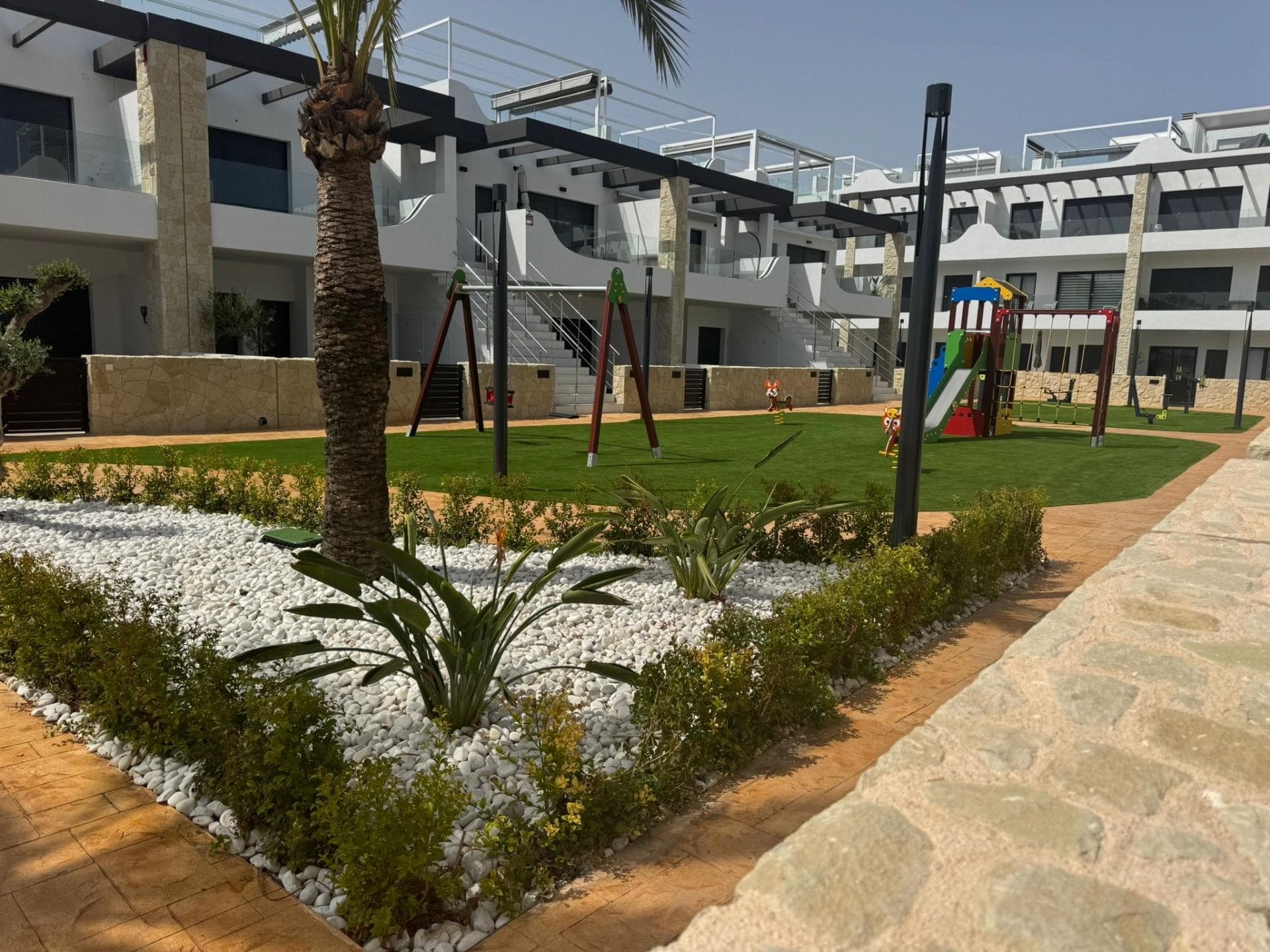 Wake up to soft Mediterranean light spilling onto your terrace, slip down to the communal pool before breakfast, and stroll along the sand at Punta Prima before lunch — this is everyday life in this beautifully turn-key ground floor apartment in one of Orihuela Costa's most loved corners. Set just one kilometre from the beach and only minutes from La Zenia Boulevard, the apartment opens onto a generous south-facing front terrace overlooking lush communal gardens and the swimming pool. Inside, a bright living-dining room flows into a fully equipped kitchen, with three comfortable bedrooms — all with fitted wardrobes — and two modern bathrooms. A second large rear terrace gives you a quieter outdoor space for shaded morning coffees and unhurried evening dinners. Sold fully furnished and ready to move into, with air conditioning throughout and an underground garage space included. Golf courses, restaurants, supermarkets and the shops of La Zenia Boulevard are all within a 2-minute drive, while Alicante-Elche airport is just 50 km away — a brilliant lock-up-and-leave home in the sun, equally suited to year-round living, a winter escape, or a strong holiday rental investment. Property reference M1077 — viewings by appointment.