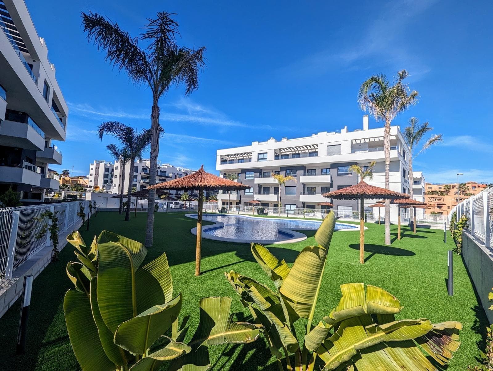 Imagine sunset drinks high above the rooftops of Villamartín, the warm orange glow of the Costa Blanca sky stretching out from your private solarium — that's the lifestyle waiting for you in this immaculate south-facing penthouse, perfectly placed between three legendary golf courses and the beaches of La Zenia. The home opens into a bright open-plan living and dining area with electric shutters and reversible hot/cold air conditioning. The fully fitted kitchen flows naturally into the lounge, and a private terrace — complete with handy utility room — extends your living space outdoors. The principal bedroom has fitted wardrobes and a stylish en-suite, while a second double bedroom with built-in storage shares a generous walk-in shower bathroom. Upstairs, the showpiece: a large rooftop solarium for sun-drenched mornings, alfresco dinners, and evenings under the stars. Residents enjoy a swimming pool, manicured green areas, petanque, a children's playground and a communal park — all just a short stroll from Villamartín Plaza with its bars, restaurants and shops. The apartment is sold furnished and includes a private parking space and lift access. Property reference M1078 — viewings by appointment.